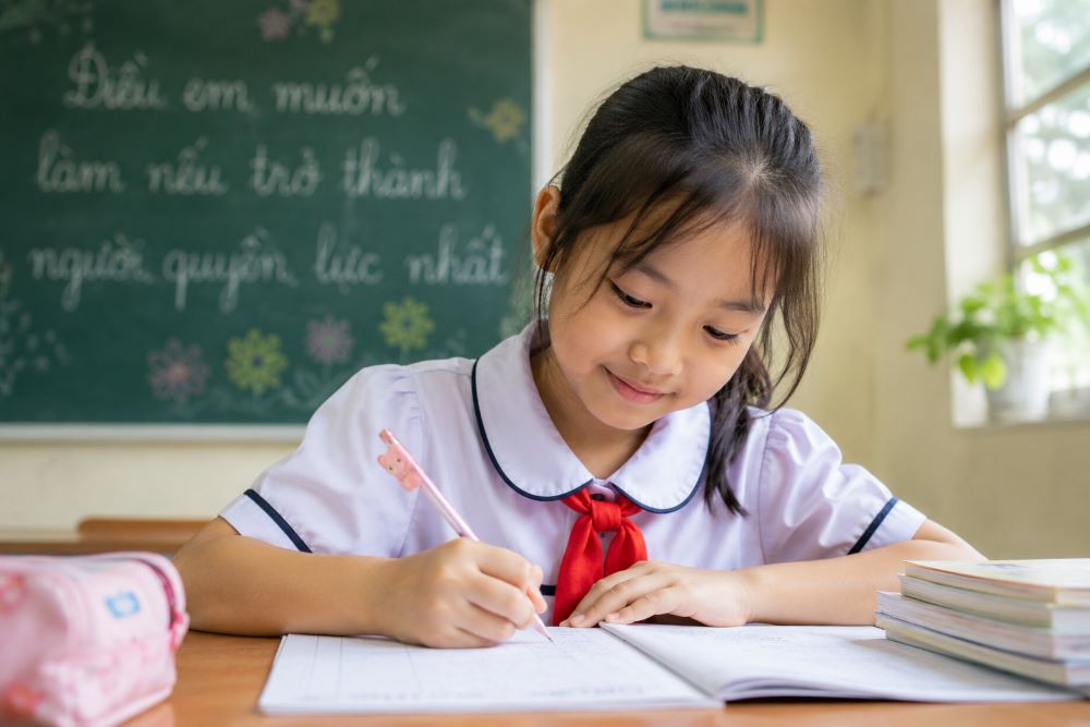 Hình ảnh cô bé lớp 5 hồn nhiên chăm chú viết bài trong lớp học, gợi liên tưởng đến câu chuyện về bài văn gây chú ý.
